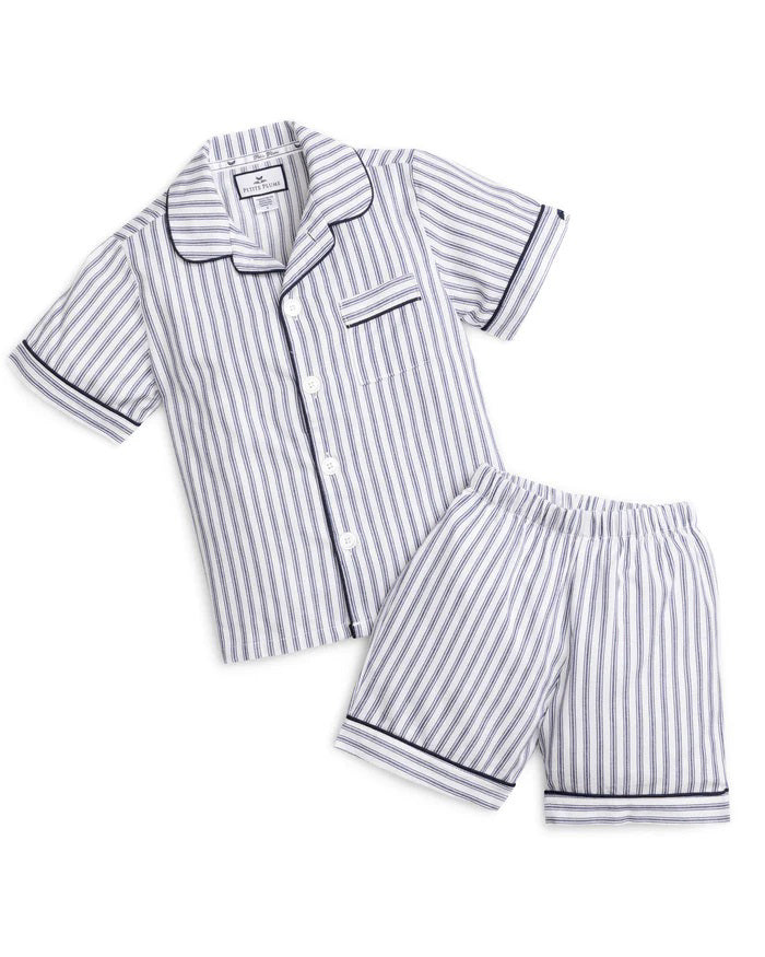 A young boy stands barefoot against a plain background, smiling and looking off to the side. He is wearing an elegant PETITE PLUME FEARRINGTON LIFESTYLE COLLECTION CHILDREN'S TWILL NAVY FRENCH TICKING PAJAMA SHORT SET and is holding a plush grey teddy bear by its arm.