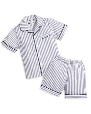 A young boy stands barefoot against a plain background, smiling and looking off to the side. He is wearing an elegant PETITE PLUME FEARRINGTON LIFESTYLE COLLECTION CHILDREN'S TWILL NAVY FRENCH TICKING PAJAMA SHORT SET and is holding a plush grey teddy bear by its arm.
