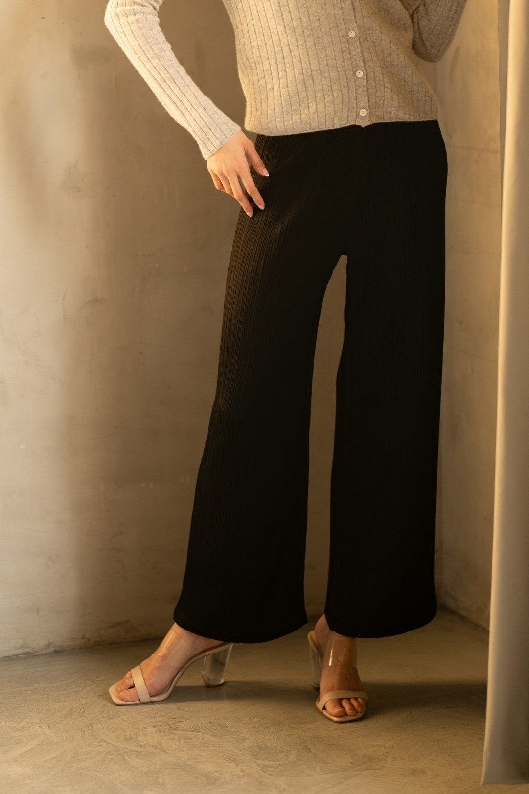 A woman smiles against a white backdrop, wearing a black top and ROSE AND CROWN - SATIN RIBBED PANT. She accessorizes with layered necklaces and a large black handbag, posing with one leg slightly raised for a relaxed look.