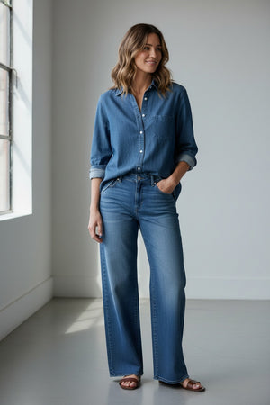 A person with long brown hair stands outdoors, facing away, next to a black wooden fence with trees and grass in the background, wearing the FRANK & EILEEN EILEEN Relaxed Button Up Shirt in Famous Denim Distressed Indigo Wash.