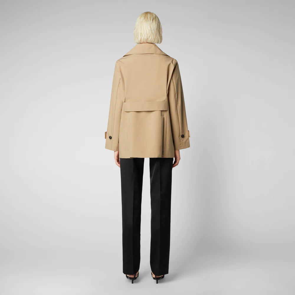 An individual with short blonde hair is wearing the SAVE THE DUCK - SOFI TRENCH JACKET, a beige, weather-resistant double-breasted coat. They are also dressed in a white top, black pants, and black pointed shoes. They are standing against a plain gray background, facing forward with arms at their sides.