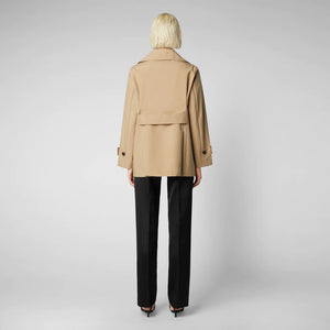 An individual with short blonde hair is wearing the SAVE THE DUCK - SOFI TRENCH JACKET, a beige, weather-resistant double-breasted coat. They are also dressed in a white top, black pants, and black pointed shoes. They are standing against a plain gray background, facing forward with arms at their sides.