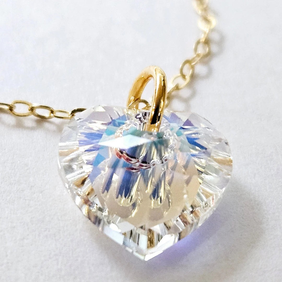 The VERDIER - AURORA CRYSTAL HEART NECKLACE from VERDIER features a sparkling crystal AB heart-shaped pendant suspended on a delicate 14k gold filled chain, shimmering against a white background.