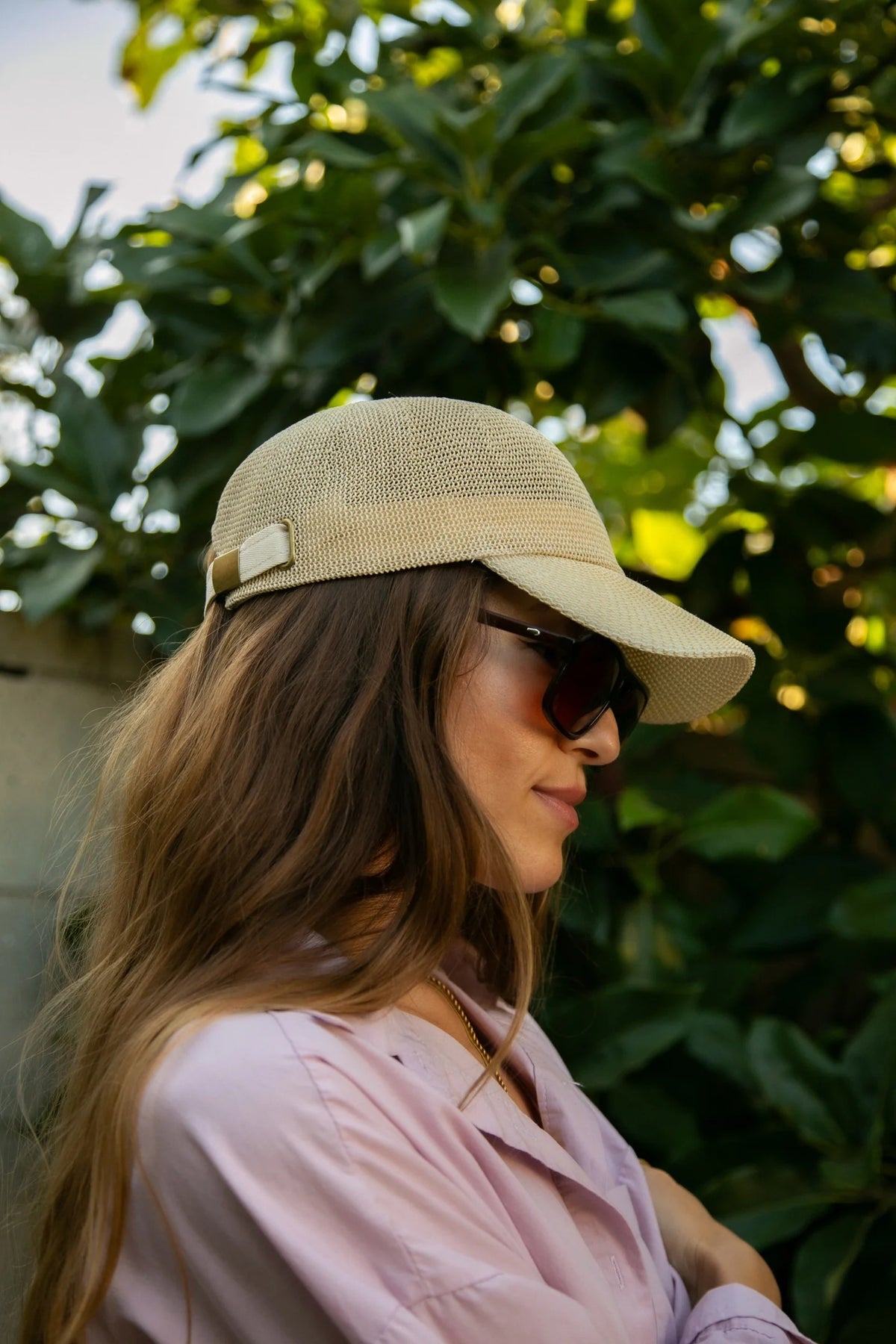 A woman with long, wavy blonde hair wears the WYETH HATS - PAULA MESH CAP IN NATURAL by WYETH and a white tank top, sitting against a plain light background and looking to the left with a neutral expression.