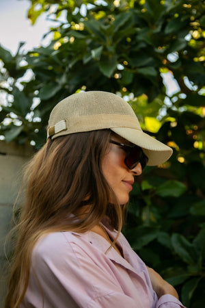 A woman with long, wavy blonde hair wears the WYETH HATS - PAULA MESH CAP IN NATURAL by WYETH and a white tank top, sitting against a plain light background and looking to the left with a neutral expression.