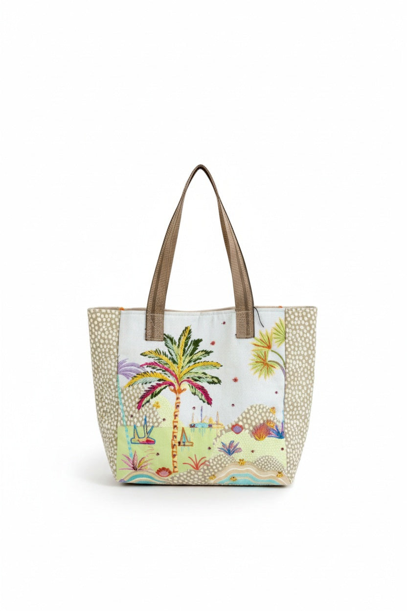 A person in pink pants, a colorful scarf, green shoes, and a light shirt holds the STORIATIPIC - CALI TOTE BAG by STORIATIPIC, an embroidered cotton bag with a palm tree and beach scene. The image is cropped above the shoulders.