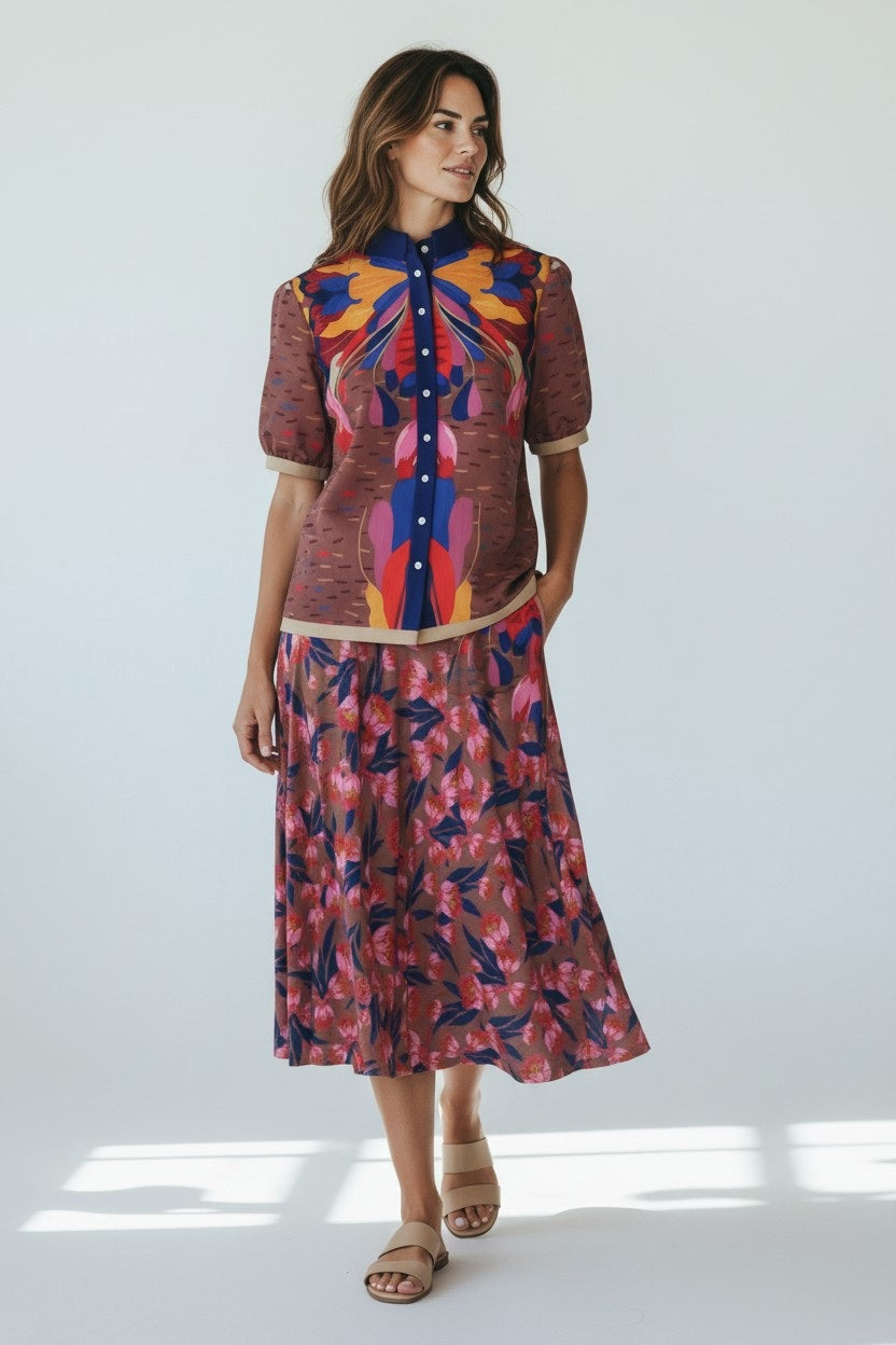 A woman with wavy hair smiles softly in a bright room, wearing a white V-neck blouse, brown strappy sandals, and the FRANCO FERRARI - FLORAL PRINT SKIRT—a high-waisted pink and navy made-in-Italy piece by FRANCO FERRARI.