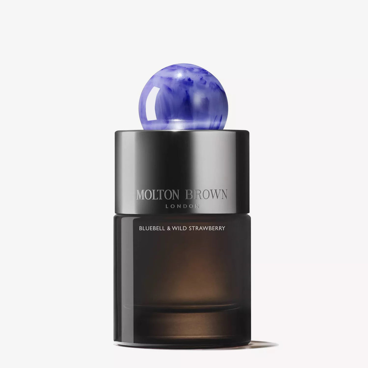 Three Molton Brown Bluebell &amp; Wild Strawberry Eau de Parfum bottles sit on a blue surface with fresh strawberries, emphasizing the collection’s wild strawberry and floral-fruity scent. A silver tube is placed on the right side.