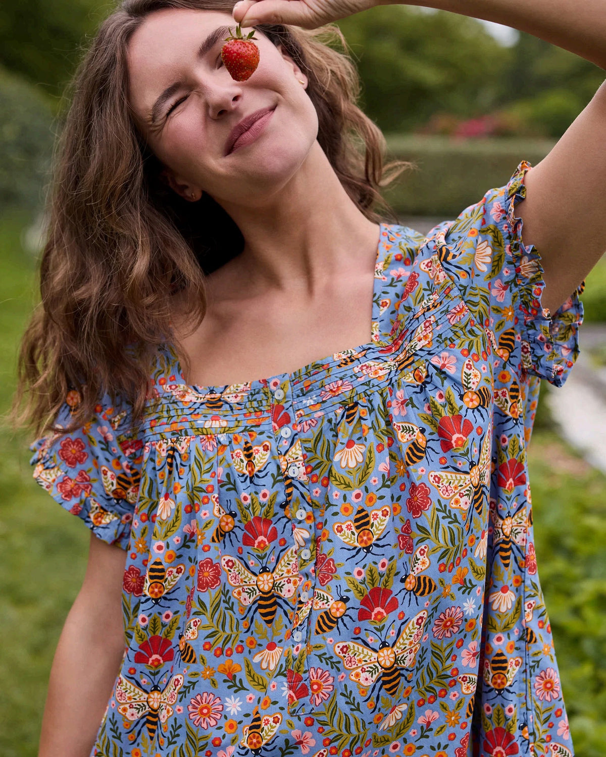 A woman with long blonde hair wears the PRINTFRESH Bee Tapestry Pinktuck Nightgown in Delphinium Blue, standing against a plain light-colored wall and smiling slightly.