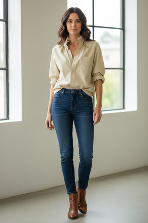 A woman in the KMJ - Southern Lover Shirt with a ruffle collar and blue jeans, featuring the iconic KMJ tuck, stands confidently beside a horse, holding its reins amidst green foliage. She's elegantly posed with jewelry, embodying Southern-inspired charm.