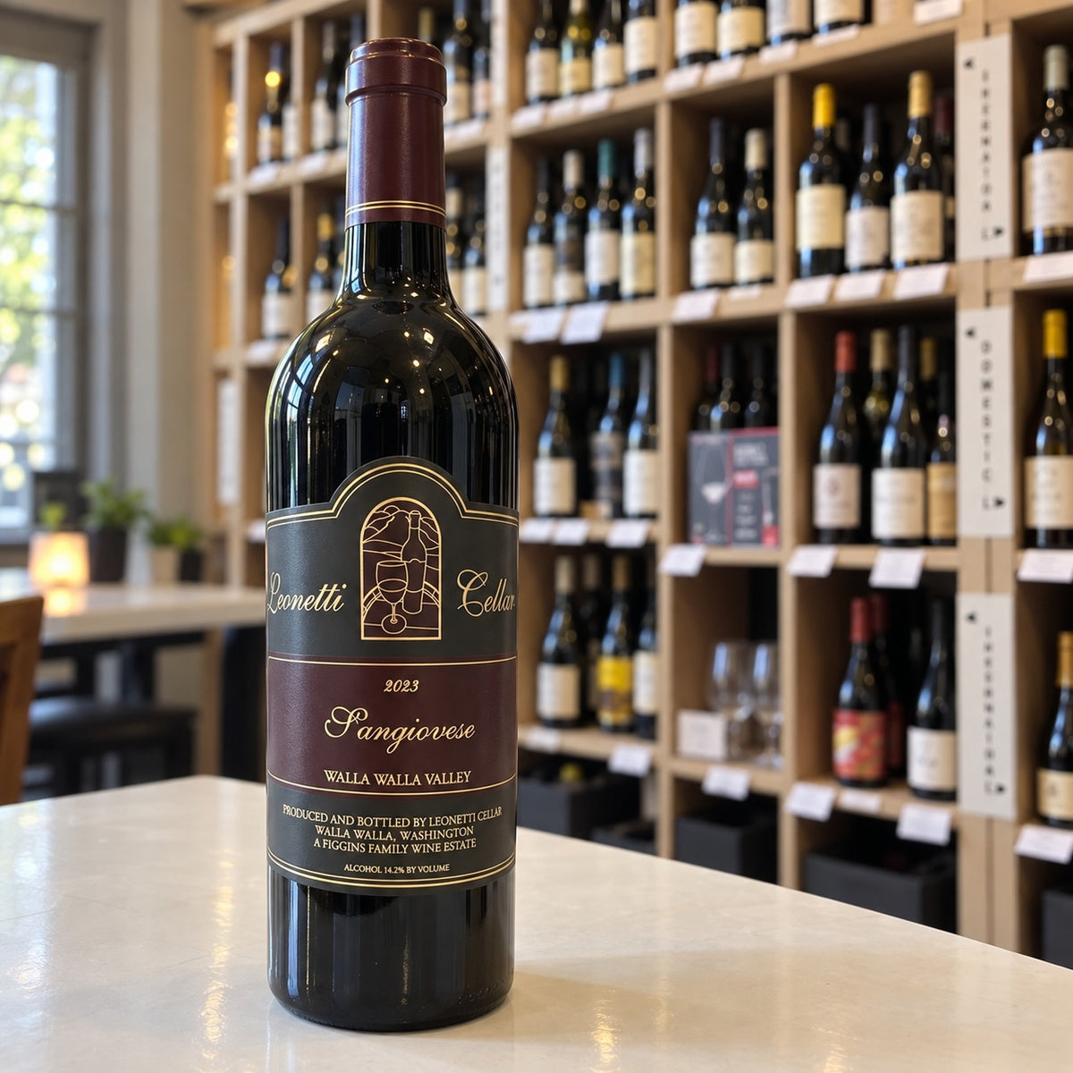 A bottle of LEONETTI SANGIOVESE 2023 by WINE: WINEBOW rests on a countertop, while shelves filled with assorted wine bottles appear blurred in the background.
