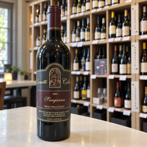 A bottle of LEONETTI SANGIOVESE 2023 by WINE: WINEBOW rests on a countertop, while shelves filled with assorted wine bottles appear blurred in the background.