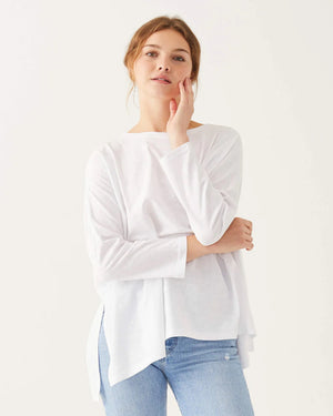 A woman with wavy brown hair models the MERSEA - CATALINA SLUB TEE—an oversized, long-sleeved white top with thin gray stripes—by MERSEA. She pairs it with blue jeans and stands against a plain, light background, hands behind her back.