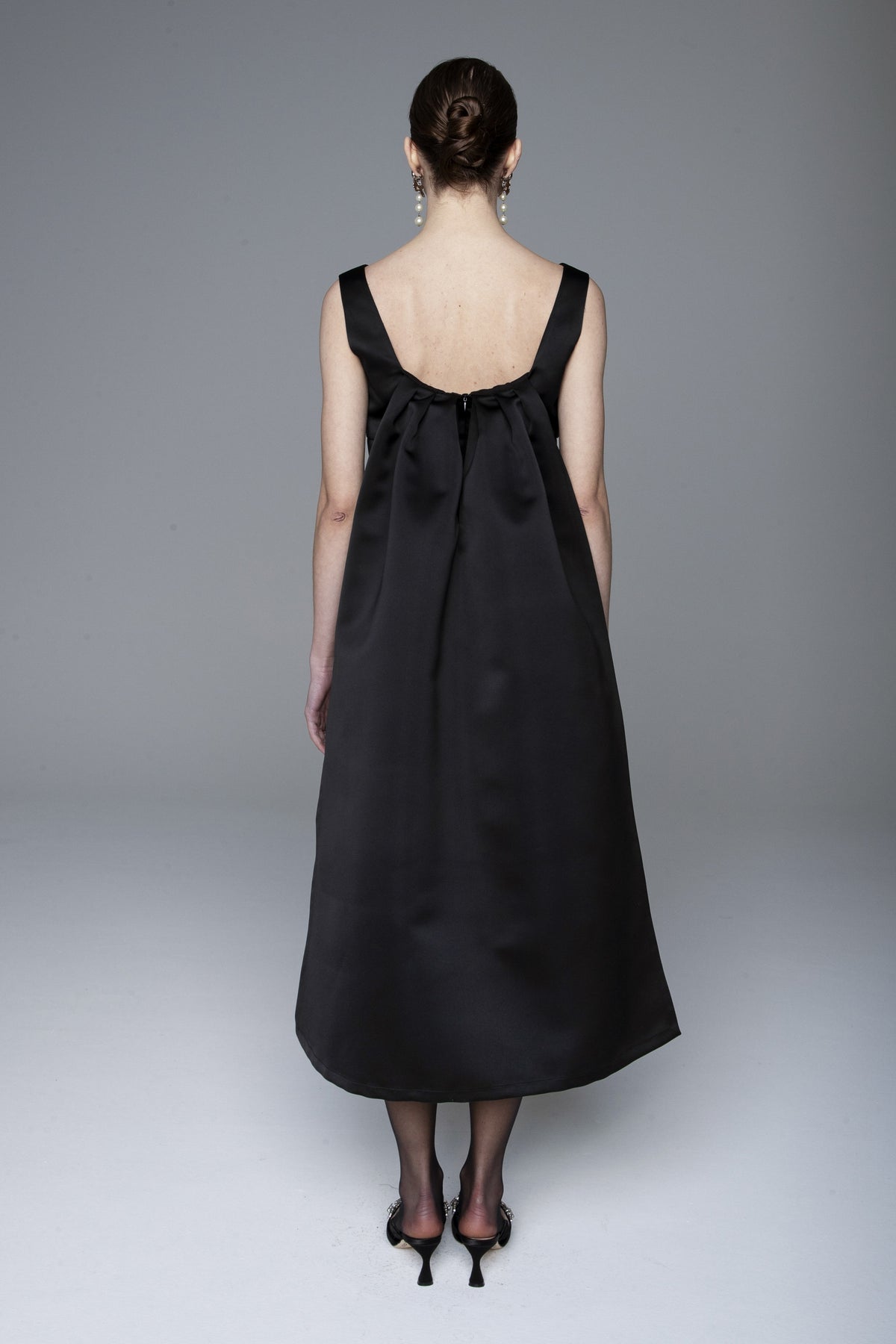 A woman stands facing away in the LAVI - NARA DRESS, a sleeveless black midi dress by LAVI reaching mid-calf. Her hair is in a neat bun, and she pairs it with black heels and drop earrings against a plain grey background.