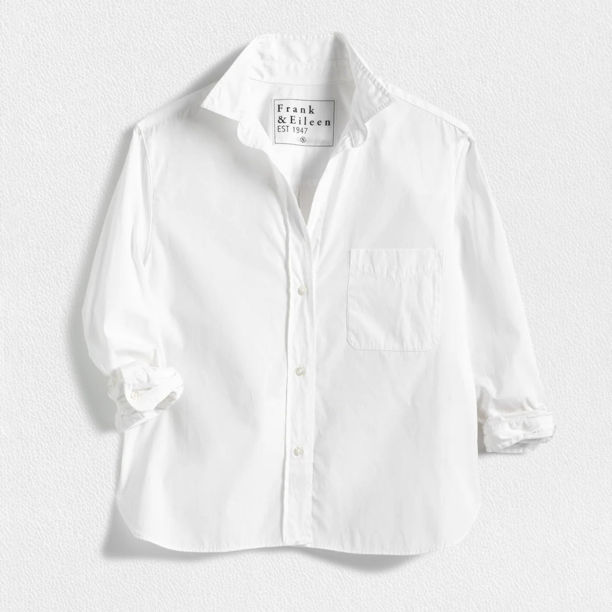 The FRANK &amp; EILEEN Silvio Untuckable Button-Up Shirt in Superluxe Cotton, featuring a chest pocket and rolled-up sleeves, is laid flat on a textured surface. The inside label reads "Frank &amp; Eileen EST 1947.