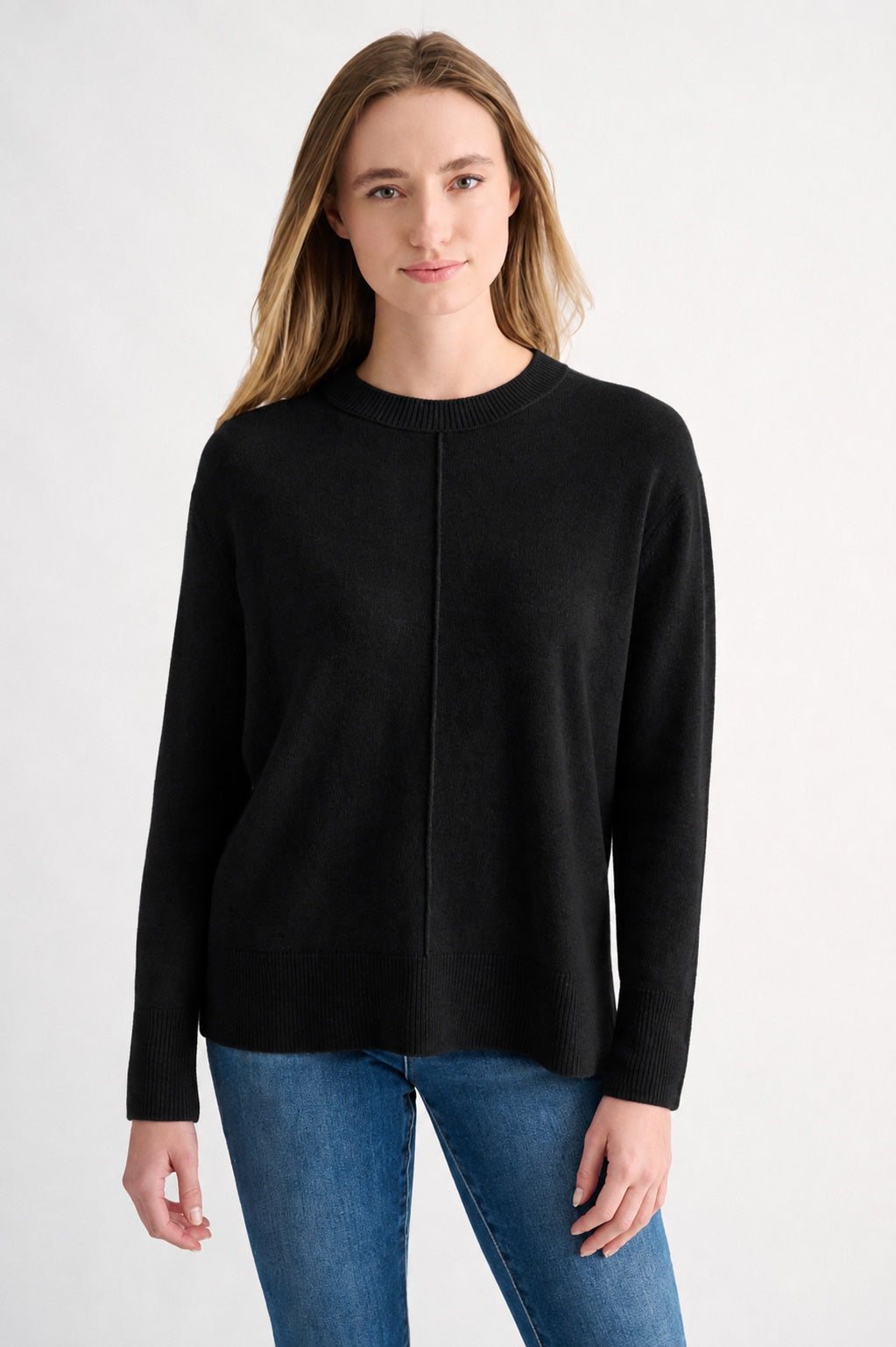 A person with long, light brown hair is wearing the KINROSS CASHMERE - HI LOW SEAMED CREW NECK CASHMERE SWEATER IN BLACK by KINROSS, styled in a relaxed classic fit with blue jeans. They are standing against a plain white background, facing the camera and smiling slightly.