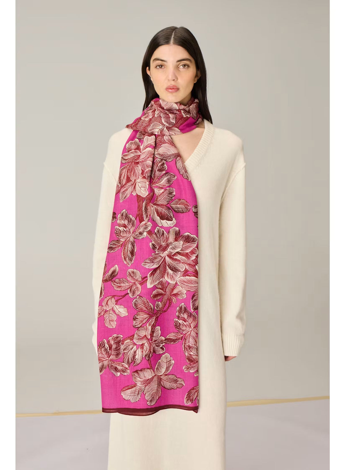A person stands against a plain beige background wearing a long cream-colored sweater dress and the INOUI EDITIONS - ARBORIS WOOL SCARF IN PINK by INOUI, a bright pink, 100% wool scarf featuring large floral patterns and made in India.