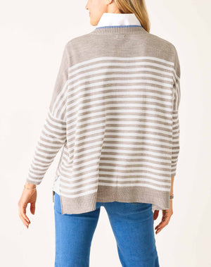 A woman with blonde hair wears the MERSEA - CATALINA MODERN CREWNECK SWEATER in gray and white stripes over a white collared shirt and blue jeans, standing against a plain white wall with a slight smile, one arm bent across her body.