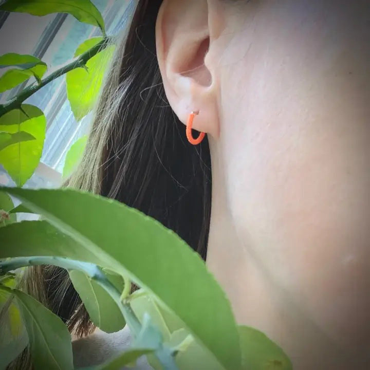 A close-up of a person’s ear wearing ALICE RISE - TINY HUGGIE EARRINGS IN NAVY, the handmade jewelry partly hidden by green leaves as natural light from a window highlights the unique ALICE RISE design.