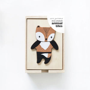 A wooden toy set titled "WEE GALLERY - MIX & MATCH ANIMAL TILES" by WEE GALLERY shown on a white background. The visible toy piece features a cartoonish animal, combining traits of a fox and a bear, painted in brown, white, and black colors. Perfect for creative play and developing motor skills, the toy is presented in a wooden box.