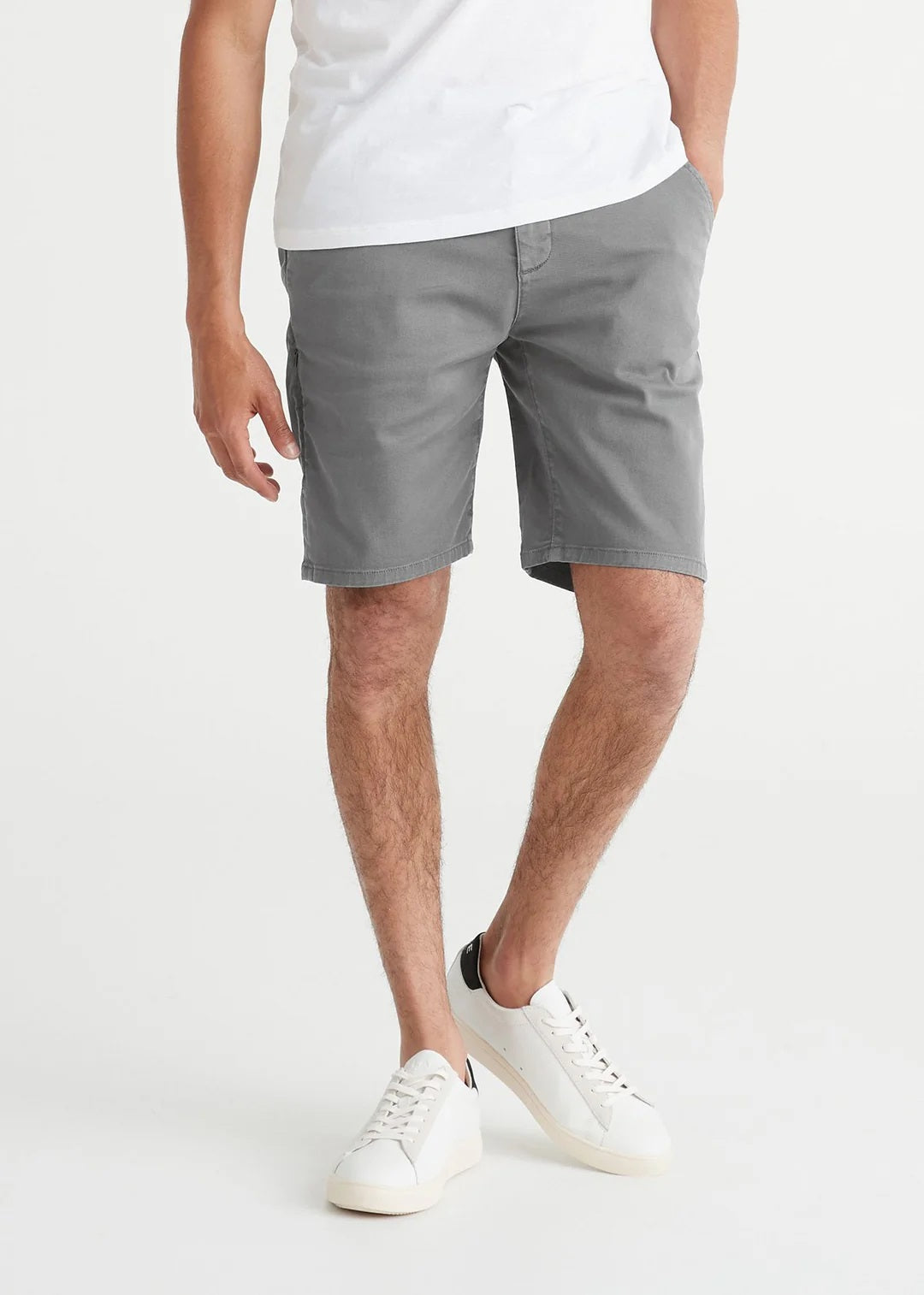 A person faces away from the camera, wearing a white T-shirt, DUER Live Free Journey Shorts in Titanium (slim fit), and white sneakers with black heel tabs, set against a plain white background.