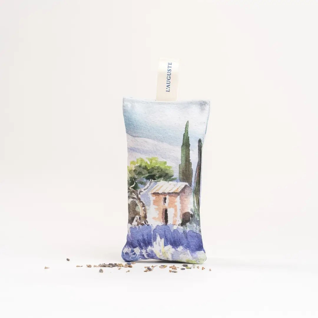 The L&#39;AUGUSTE PROVENCE - FRENCH LAVENDER SACHET FROM PROVENCE is a small handcrafted pouch with a countryside watercolor design, upright shape, &quot;L&#39;AUGUSTE PROVENCE&quot; tag, and dried lavender pieces at its base.