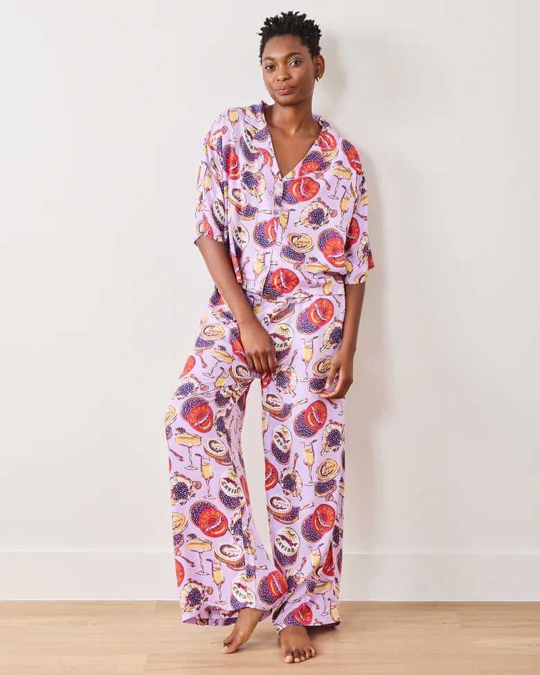 A person stands barefoot against a white wall, wearing the PRINTFRESH - ECO SATIN CHAMPAGNE AND CAVIAR PAJAMA SET by PRINTFRESH. The set features a loose fit, playful fruit print in light purple, and the person appears relaxed and confident with short hair.