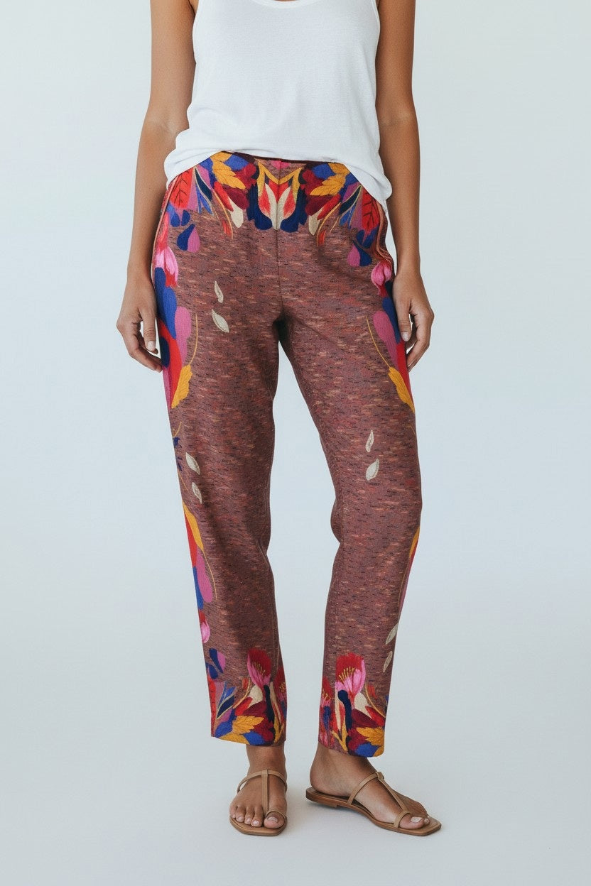 A person stands indoors wearing FRANCO FERRARI - SILK PRINT TROUSER, featuring loose-fitting silk with colorful abstract and subtle floral designs in brown, pink, and blue. They pair it with gold shoes, posed before sheer draped fabric and wooden objects.