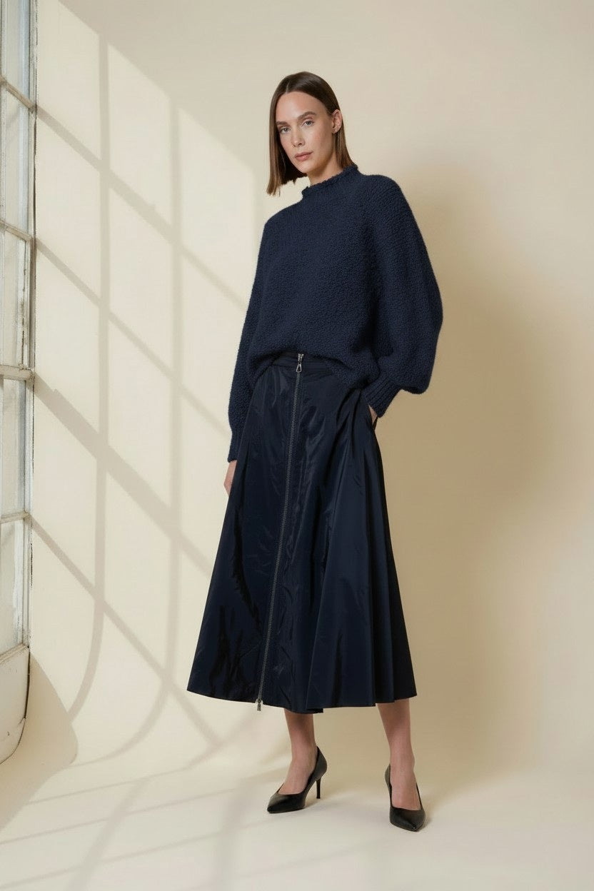 A woman with straight brown hair wears the VICARIO CINQUE MADEIRA Boucle Sweater in midnight blue, paired with a shiny navy skirt, standing against a beige wall dappled with window shadows.