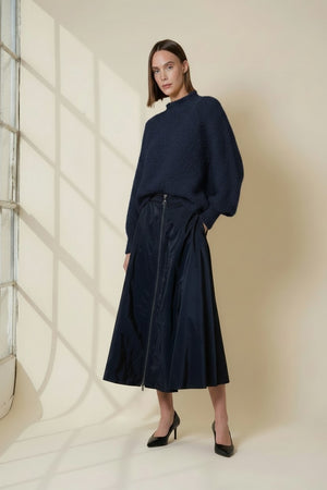 A woman with straight brown hair wears the VICARIO CINQUE MADEIRA Boucle Sweater in midnight blue, paired with a shiny navy skirt, standing against a beige wall dappled with window shadows.