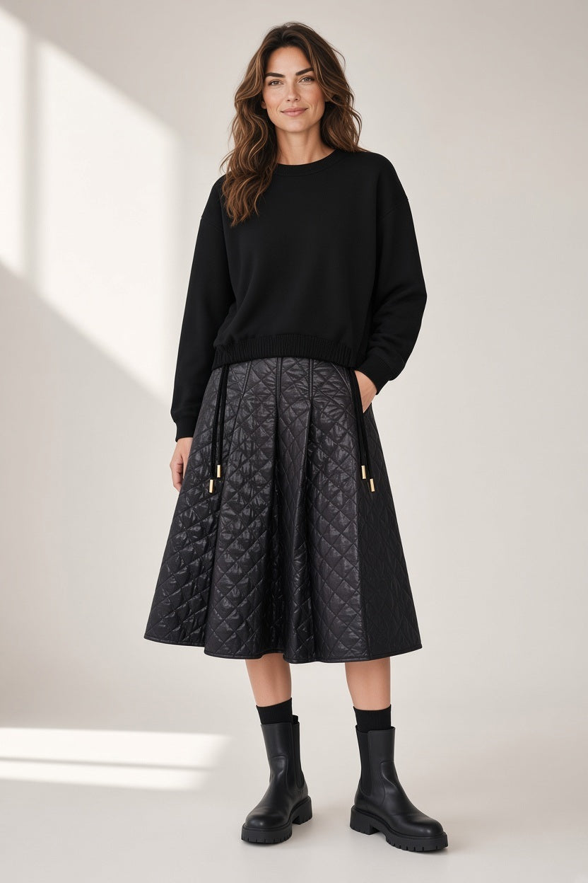 A woman wearing a black SAVE THE DUCK - AKEMI SWEATER IN BLACK crewneck sweater, a black quilted midi skirt, and black ankle boots.