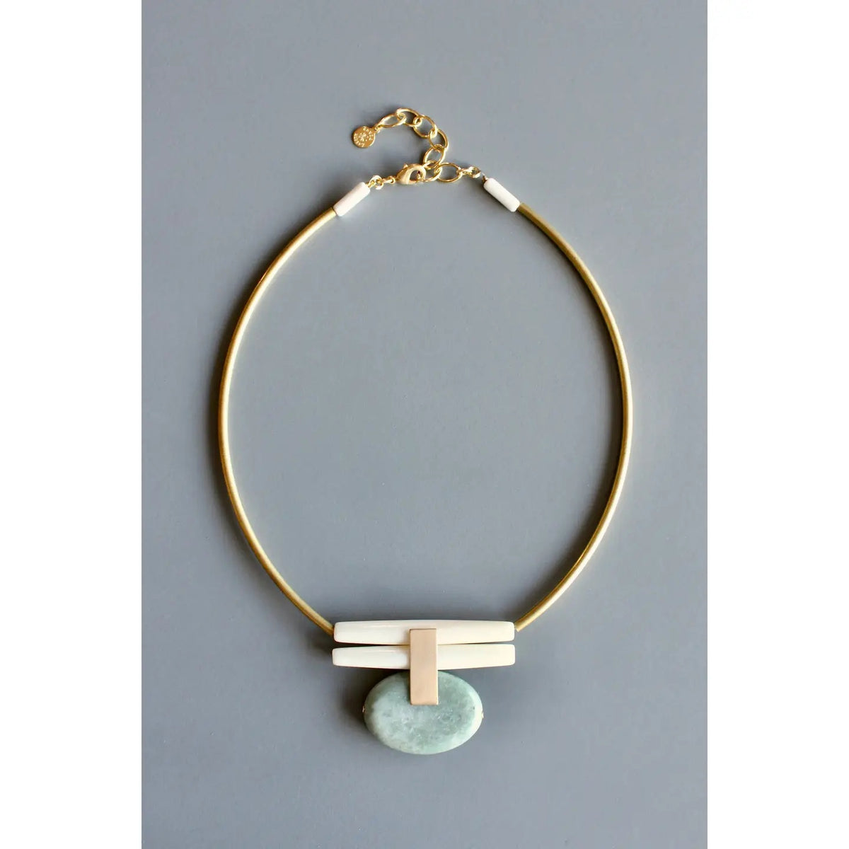 The DAVID AUBREY - GREEN JASPER AND BRASS ARTDECO NECKLACE by DAVID AUBREY features a gold chain with a light blue oval stone pendant that evokes the unique look of teal green jasper, complemented by white rectangular accents and an adjustable chain clasp, all presented on a gray background.