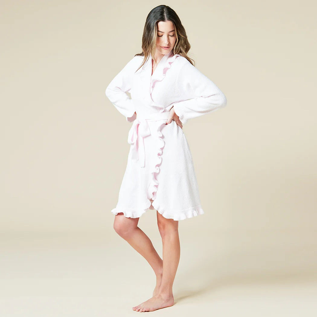 A woman with long hair stands barefoot against a light beige background, smiling softly in the SOFTIES - RUFFLE CHENILLE ROBE 36" IN PINK, featuring a belted waist and ruffled edges, exuding comfort and elegance.