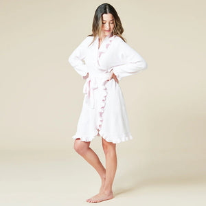 A woman with long hair stands barefoot against a light beige background, smiling softly in the SOFTIES - RUFFLE CHENILLE ROBE 36" IN PINK, featuring a belted waist and ruffled edges, exuding comfort and elegance.