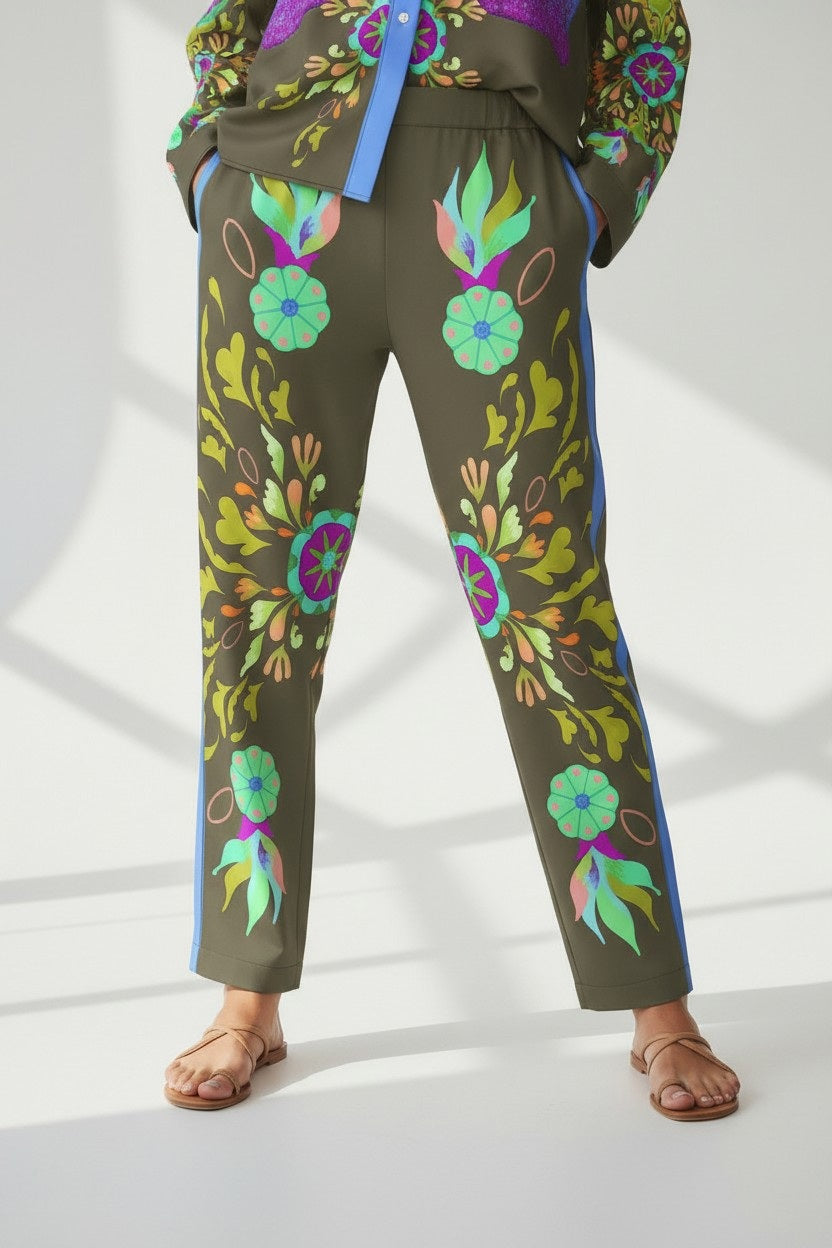 A person in FRANCO FERRARI SILK PRINT TROUSER—Italian-made, olive green with colorful florals and blue side stripes—matched with a coordinated top and tan sandals, stands against a bright background with soft shadows.
