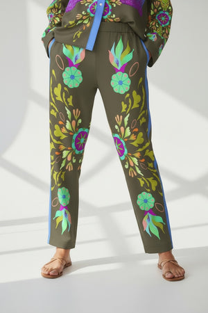 A person in FRANCO FERRARI SILK PRINT TROUSER—Italian-made, olive green with colorful florals and blue side stripes—matched with a coordinated top and tan sandals, stands against a bright background with soft shadows.
