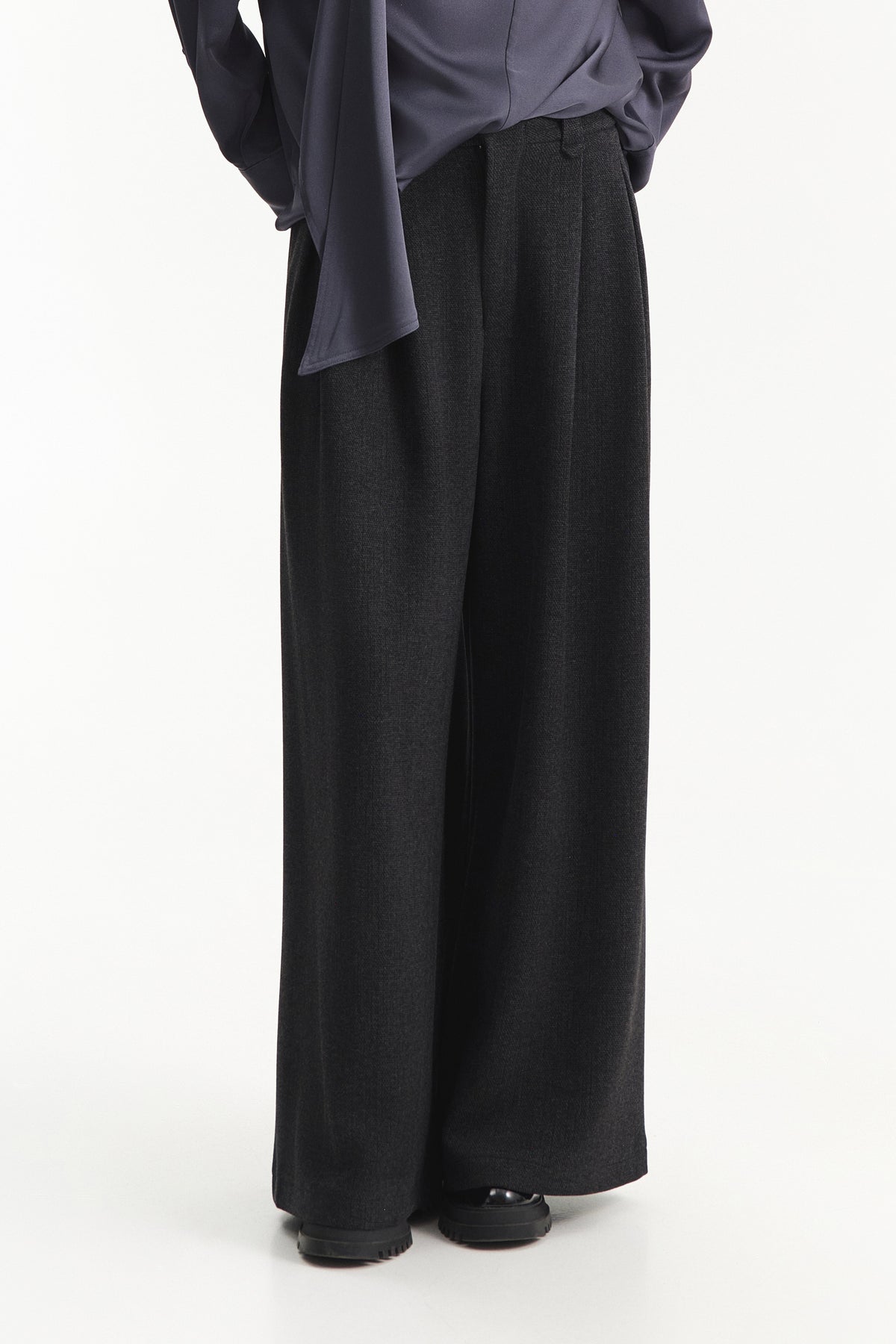 A person is shown from the shoulders down against a plain white background, wearing ELEH - BLACK PALAZZO TROUSERS by ELEH styled with black shoes and a loose-fitting, long-sleeve top.
