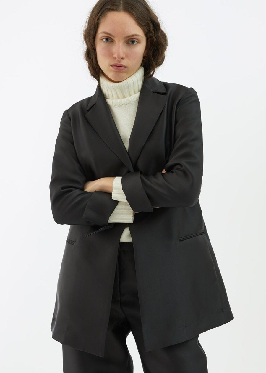 A person with long, wavy hair is wearing the BLACK BLAZER / JACKET by ALYKI LABORATORIO, made in Italy, over a white turtleneck sweater. They are standing against a white background with their arms crossed and have a serious expression on their face.