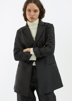 A person with long, wavy hair is wearing the BLACK BLAZER / JACKET by ALYKI LABORATORIO, made in Italy, over a white turtleneck sweater. They are standing against a white background with their arms crossed and have a serious expression on their face.