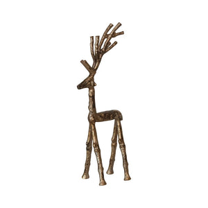 Two metallic reindeer figurines—a CREATIVE COOP CAST ALUMINUM TWIG DEER in an antique gold finish and an abstract long-legged deer—stand beside a small white bottlebrush tree on a glitter-dusted wooden surface.