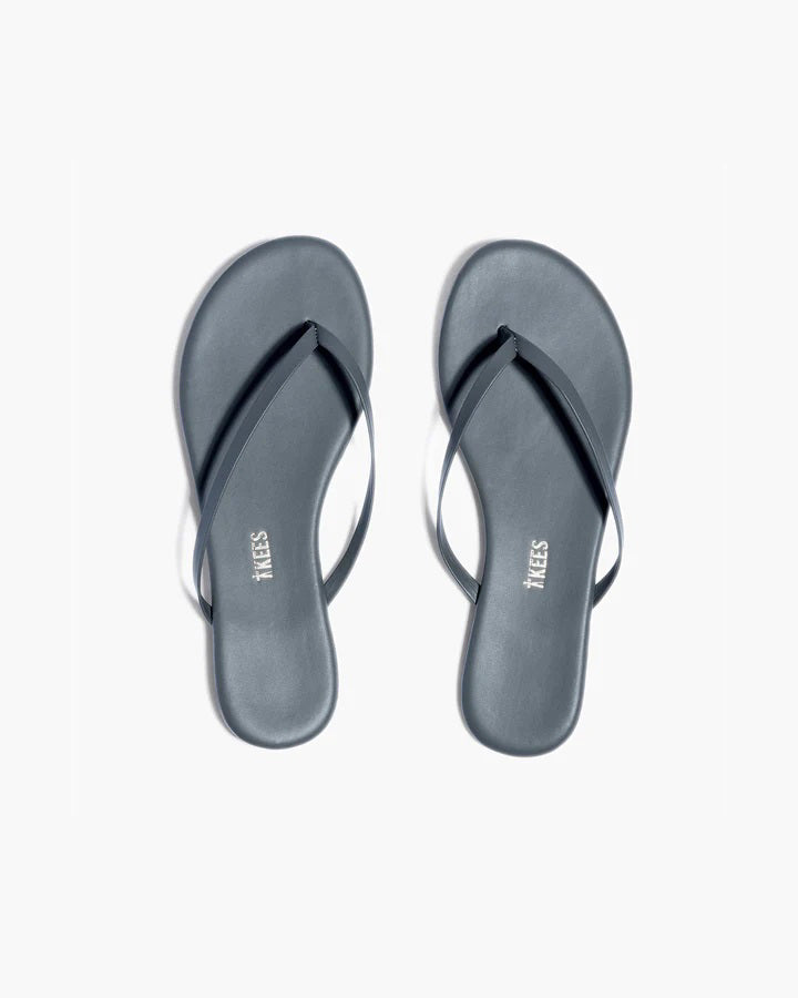 A pair of TKEES INC LILY LINER SLIPPER ASH black flip-flops with thin straps, made from Brazilian leather, are placed side by side on a plain white background with the &quot;TKEES&quot; logo visible on the cushioned insole.