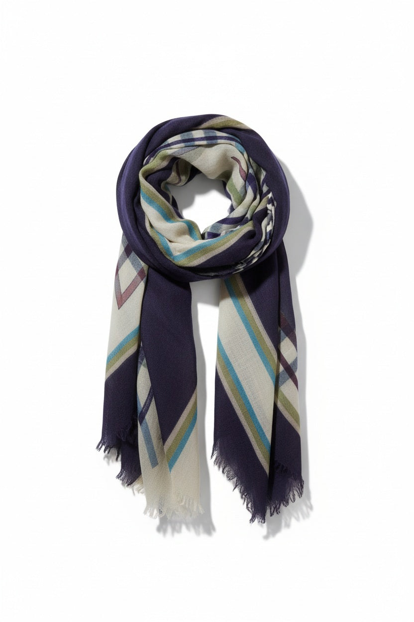 A woman in a blue shirt and jeans stands holding the STORIATIPIC - JULIE WOOL SCARF, a luxury plaid accessory in earthy tones. Behind her, mountains, beach, and grassy dunes create a scenic backdrop.