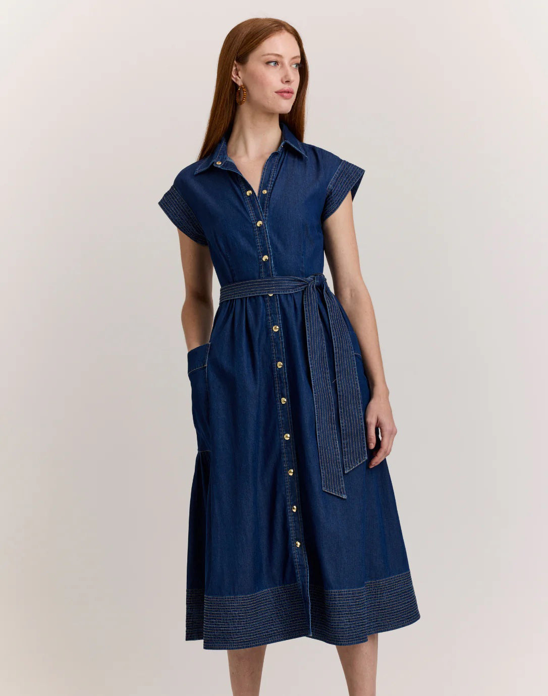 A woman stands against a light background wearing the HINSON WU Annabelle Short Sleeve Dark Wash Denim Dress, featuring a collar, button-down front, belt, pockets, and decorative stitching at the hem and sleeves.