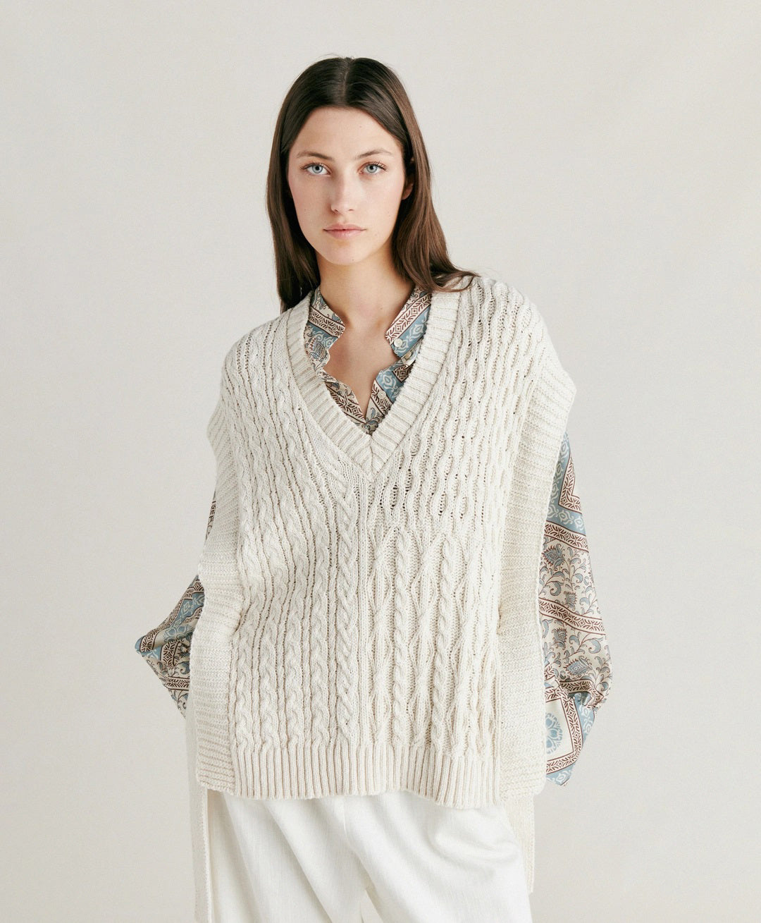 A woman with straight brown hair stands against a plain background. She is wearing the MOMONI - ARENE KNIT VEST from MOMONI, an oversized, cream-colored cable knit vest made from cotton yarn, over a patterned, long-sleeve button-up shirt and paired with white pants. Her hands are in her pockets, and she has a neutral expression.