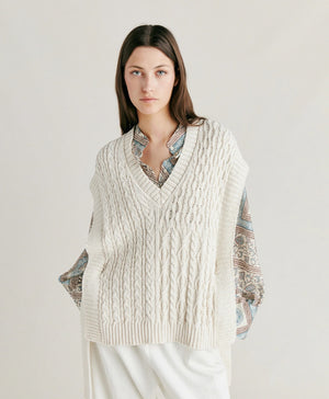A woman with straight brown hair stands against a plain background. She is wearing the MOMONI - ARENE KNIT VEST from MOMONI, an oversized, cream-colored cable knit vest made from cotton yarn, over a patterned, long-sleeve button-up shirt and paired with white pants. Her hands are in her pockets, and she has a neutral expression.