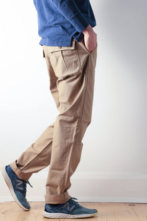 The RELWEN Supply Pant in Worker Khaki is a pair of light brown chinos inspired by classic military utility pant design, featuring side adjuster tabs, a button fly, broken twill fabric, and cuffs at the ankles with a subtly tapered leg silhouette—laid flat on a white background.
