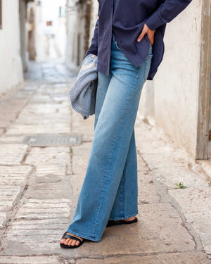 Someone stands against a neutral background wearing the MERSEA KATE WIDE LEG DENIM—light blue, high-waisted jeans made from recycled cotton—paired with a navy top, gray cardigan, and tan platform sandals.
