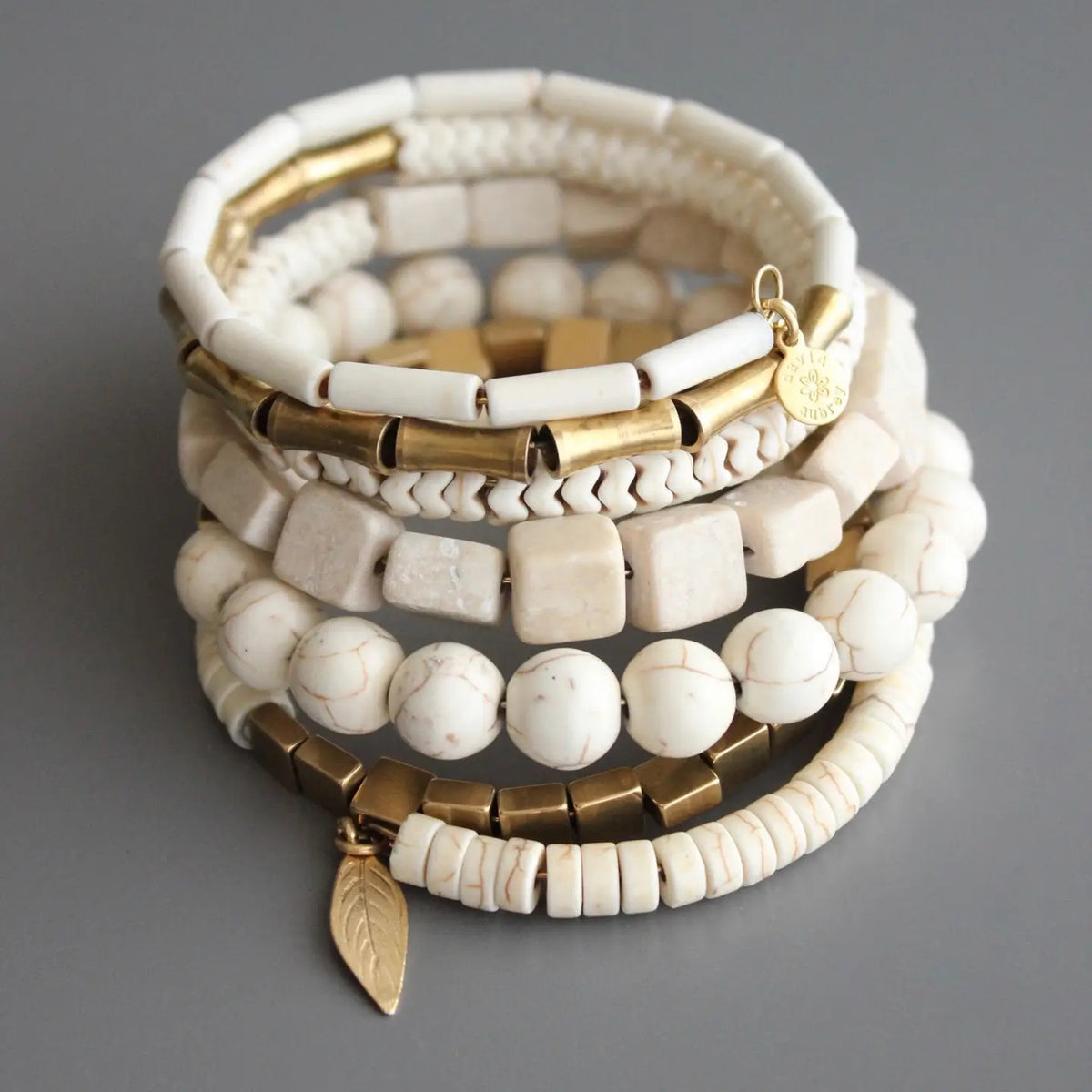 A stack of six DAVID AUBREY WHITE STONE AND BRASS WRAP BRACELETS, featuring white stones, gold beads, and brass charms—including a disc and leaf—are artfully displayed on a gray surface.