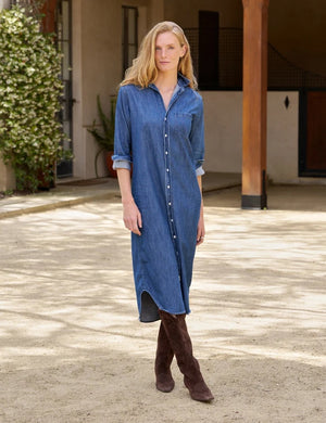 A woman stands outdoors in the FRANK & EILEEN Rory Maxi Shirtdress in Distressed Indigo Washed Denim, its long button-up silhouette and rolled sleeves crafted from luxurious indigo fabric. She pairs the effortlessly chic dress with brown knee-high boots, set against a backdrop of lush greenery and a building.