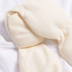 The NODPOD Weighted Sleep Eye Mask in Bone by NODPOD is a beige, segmented, plush mask with wider ends and a gently wavy, narrower middle section, shown laid flat on a white background—ideal for providing gentle pressure and soothing comfort as a weighted eye mask.